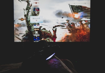 A dimly lit room with a television screen displaying a dynamic, action-packed video game interface featuring a historical battle scene. The foreground shows the silhouette of a gaming console controller held by a person.